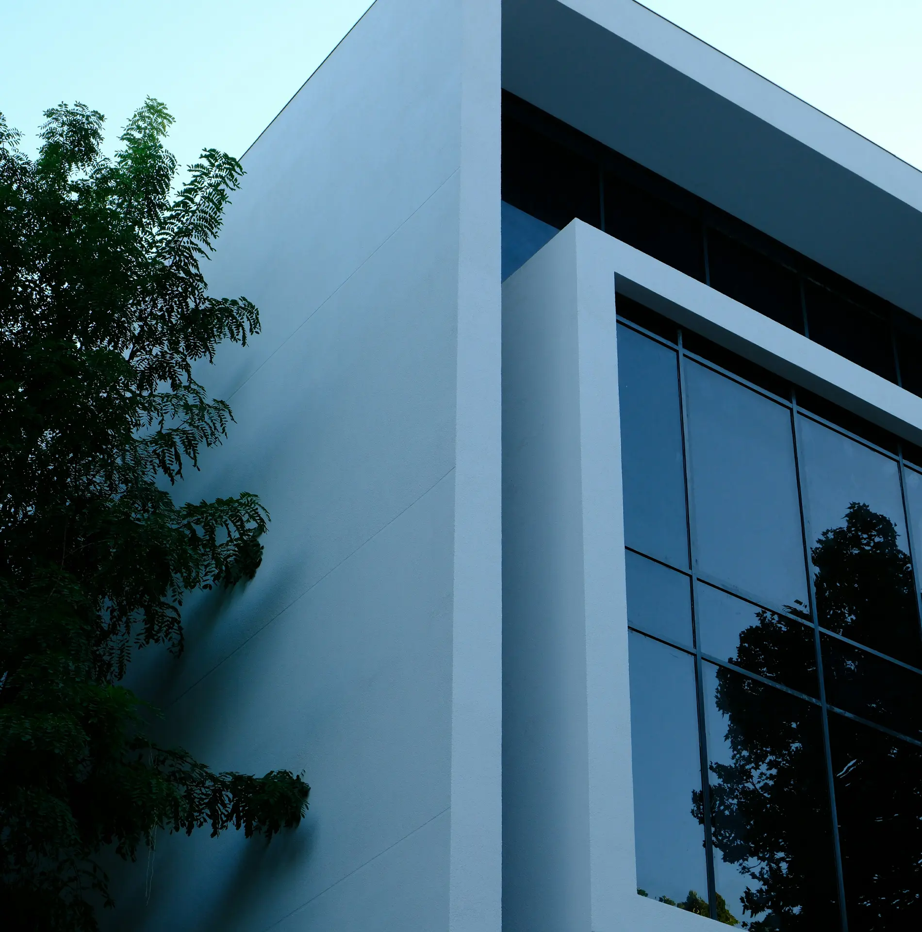 Modern architectural exterior view showing the corner of a white-rendered building with floor-to-ceiling glazing on one side, the glass reflecting nearby trees under a pale sky.
